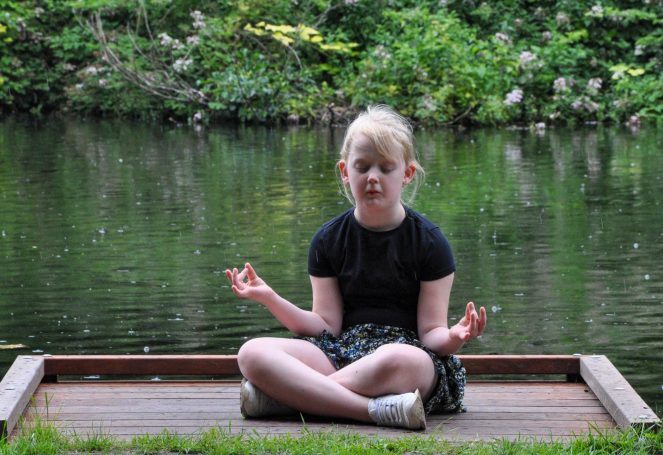 Meisje in een zwarte top en rok, mediterend aan de oever van een vijver.