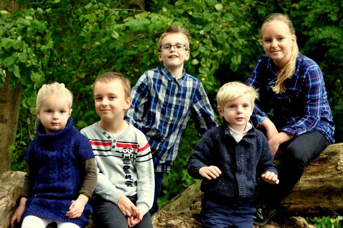 Vijf kinderen poseren samen op een boomstronk in een groene omgeving.