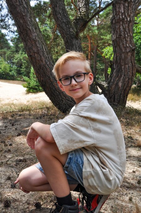 Jongen met blond haar en bril zit op de grond naast een boom in een bosrijke omgeving.