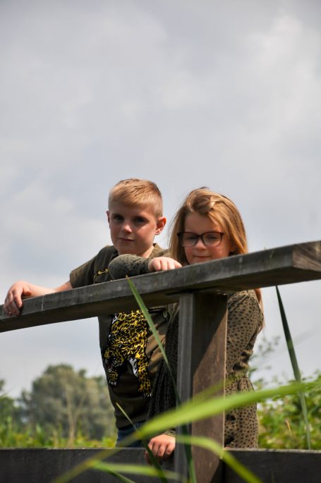 Twee kinderen staan op een houten brug met groene omgeving en een bewolkte lucht.