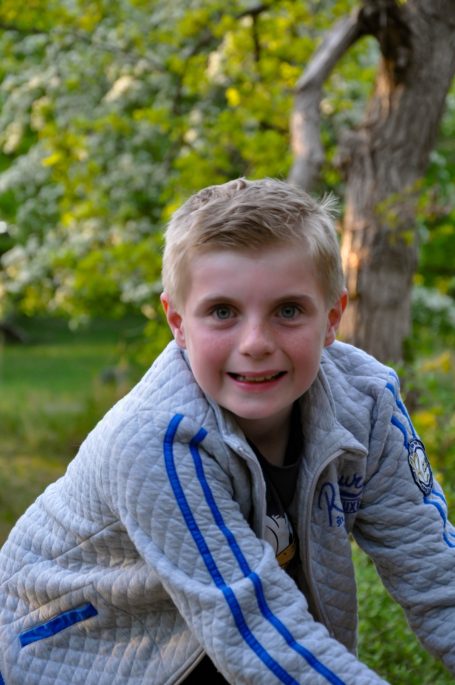 Blonde jongen in een grijze trui, glimlachend met groene bomen op de achtergrond.