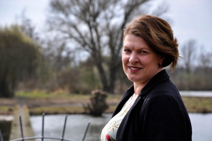 Vrouw met een glimlach, staande naast een waterpartij en bomen op de achtergrond.