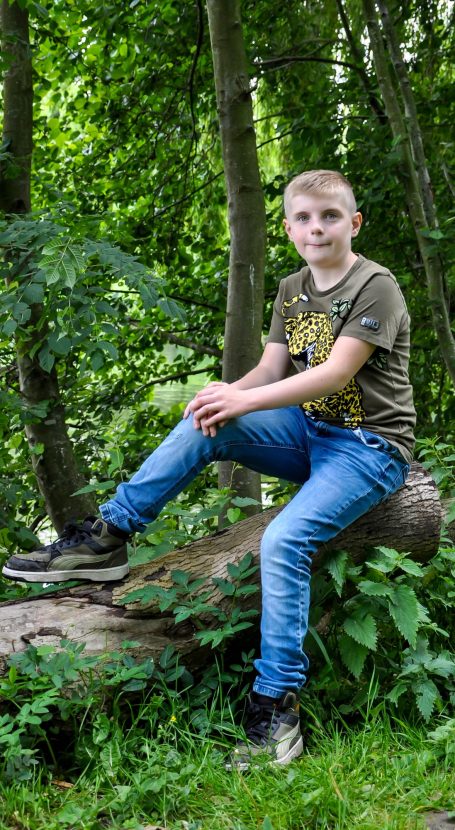 Jongen in groene omgeving, zittend op een boomstronk met een ontspannen houding.