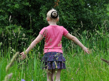 Geniet in het bos tijdens de shoot Meisje met een roze shirt en korte rok, met uitgestrekte armen in een groene grasweide.