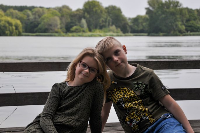 Twee kinderen zitten op een houten bankje bij het water, met groenrijke achtergrond.