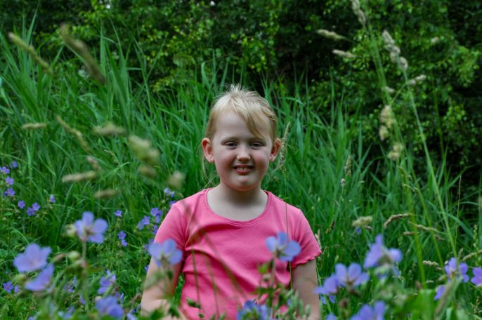 Blond meisje in een roze shirt staat tussen paarse bloemen en groene gras.