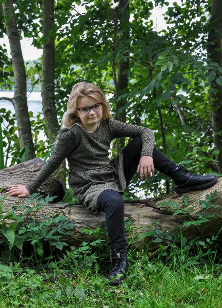 Jonge persoon met bruin haar, zittend op een boomstronk, omringd door groen.