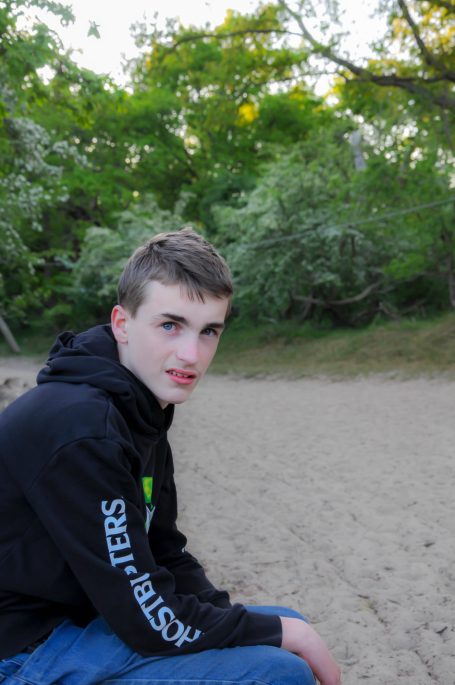 Jonge man met kort bruin haar, zittend op een zandstrand omgeven door groen.
