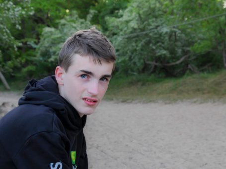 portret van een puber Jongeman met een zwarte sweater zittend op een strand met groene bomen op de achtergrond.