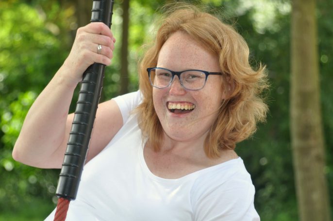Vrouw met een glimlach, holding een stok, in een groene buitenomgeving.