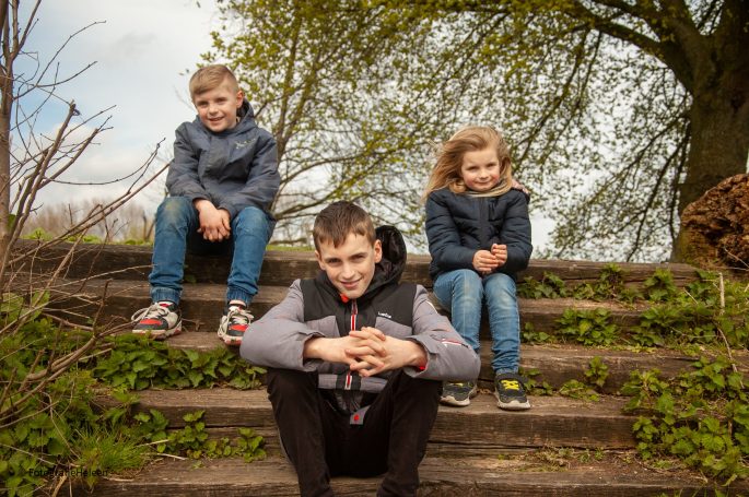 Drie kinderen zitten op trappen, omringd door groene bomen en een heldere lucht.