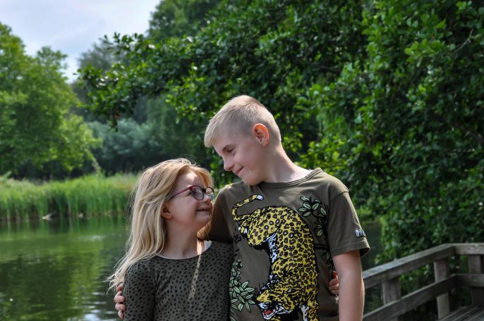 Twee kinderen staan naast elkaar bij een waterpartij, omringd door groene natuur.